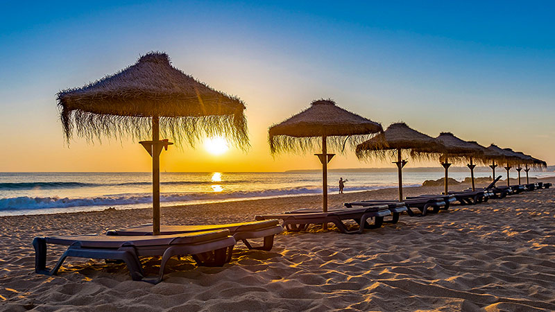 A row of tables with thatch-covered umbrellas on a sandy beach at sunset.