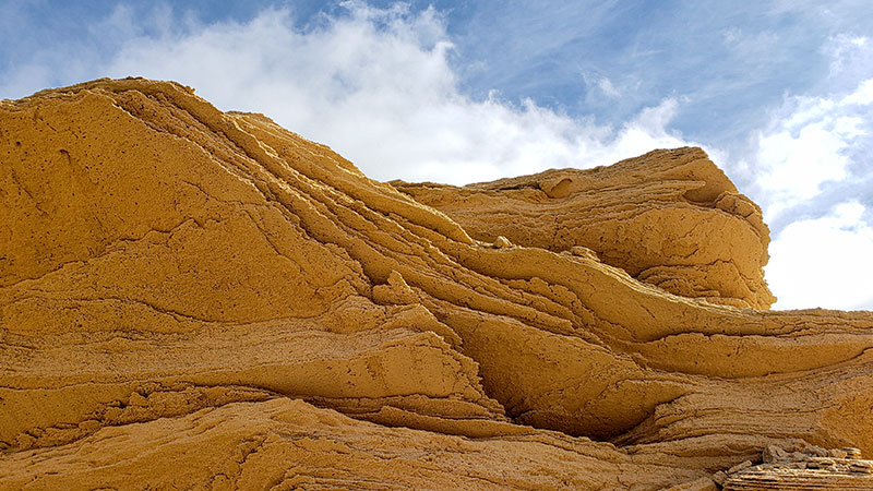 Golden layered rock against a blue sky with fluffy white clouds