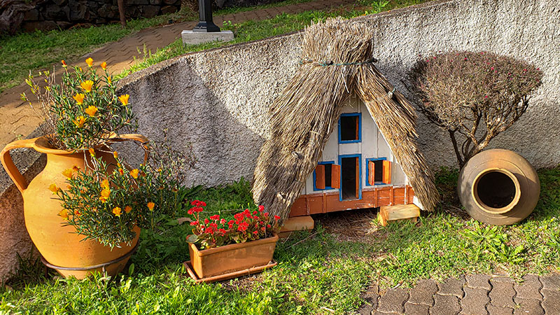 A miniature house with a grass roof and potted flowers agains a cement wall