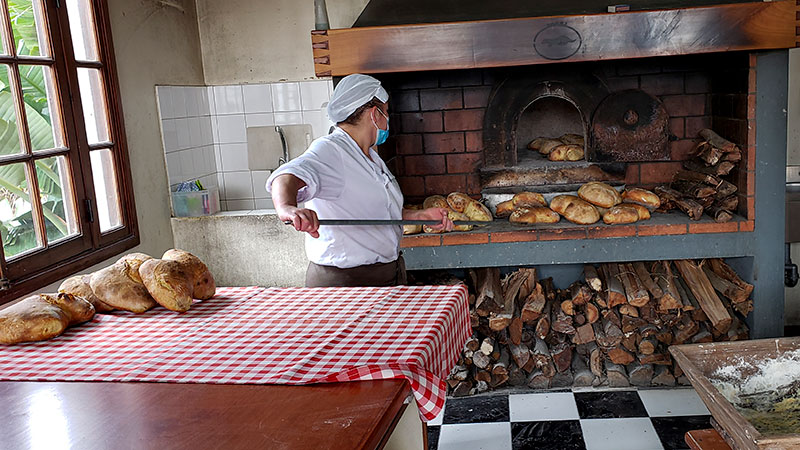 A woman uses a long-handles spatula to remove bread from a wood-fired oven.