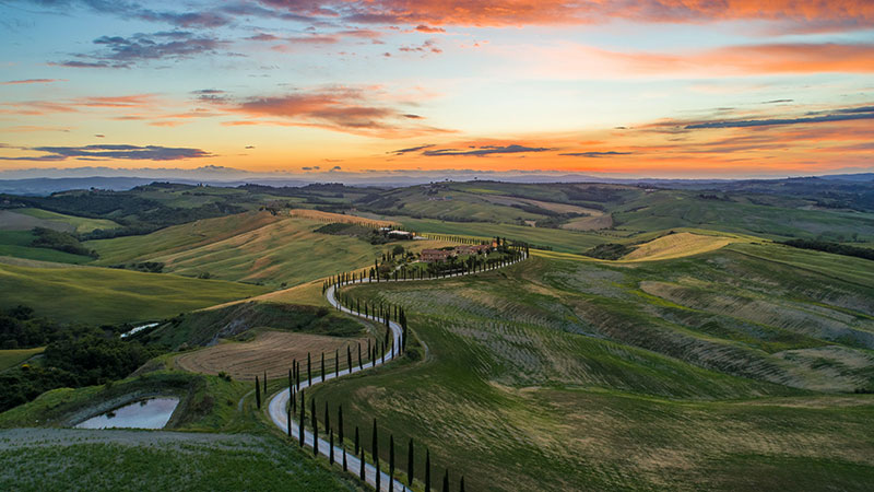 A winding road meanders through green hills in the Tuscany sunset