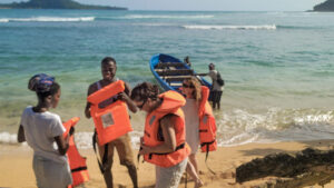 People donning bright orange life jackets on a beach, while a small boat awaits in the water.