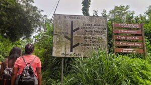 Two signs nestled amongst lush greenery, as two visitors walk along the path.