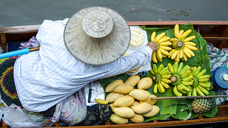 Overhead view of a canoe with a person wearing a traditional Thai hat and reaching for a bunch of bananas.